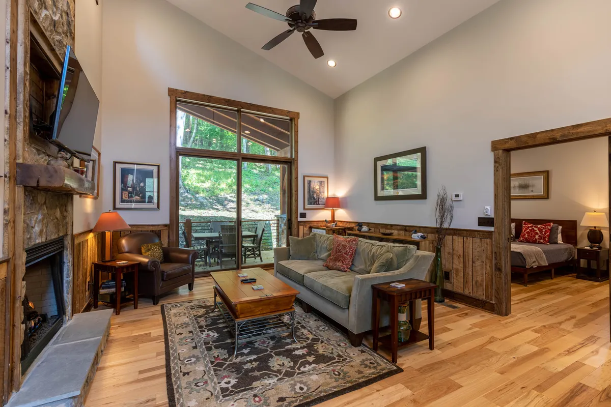 This interior living area features a striking stone fireplace, large windows that flood the room with light, a comfortable sofa with colorful cushions, and a wooden coffee table. The arrangement is ideal for relaxing, conversation, and enjoying the mountain ambiance.