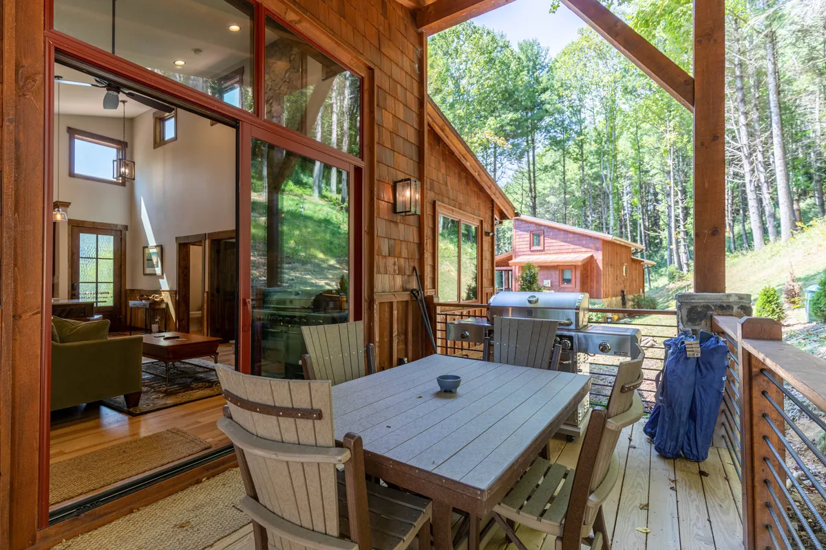 A covered outdoor deck with a dining table set for meals, surrounded by trees and featuring a barbecue grill. Large windows connect the deck to the interior, suggesting seamless indoor-outdoor living and entertaining.