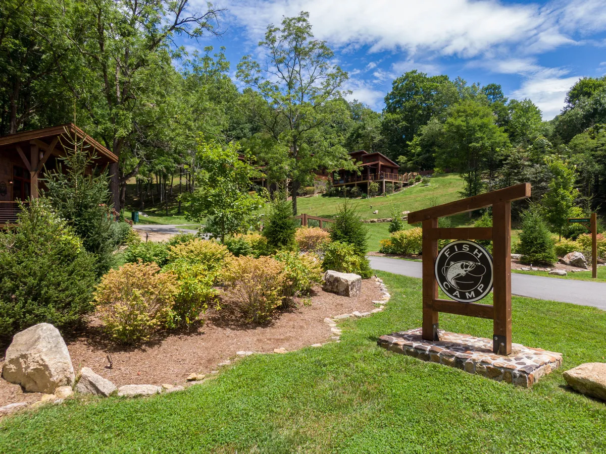 An inviting exterior scene featuring the 'Fish Camp' wooden sign, a well-maintained path, and rustic cabins nestled in lush greenery. The image hints at the peaceful fishing and nature experiences available, with clear references to fishing, hiking, and outdoor storytelling around a fire—activities central to the amenities described.