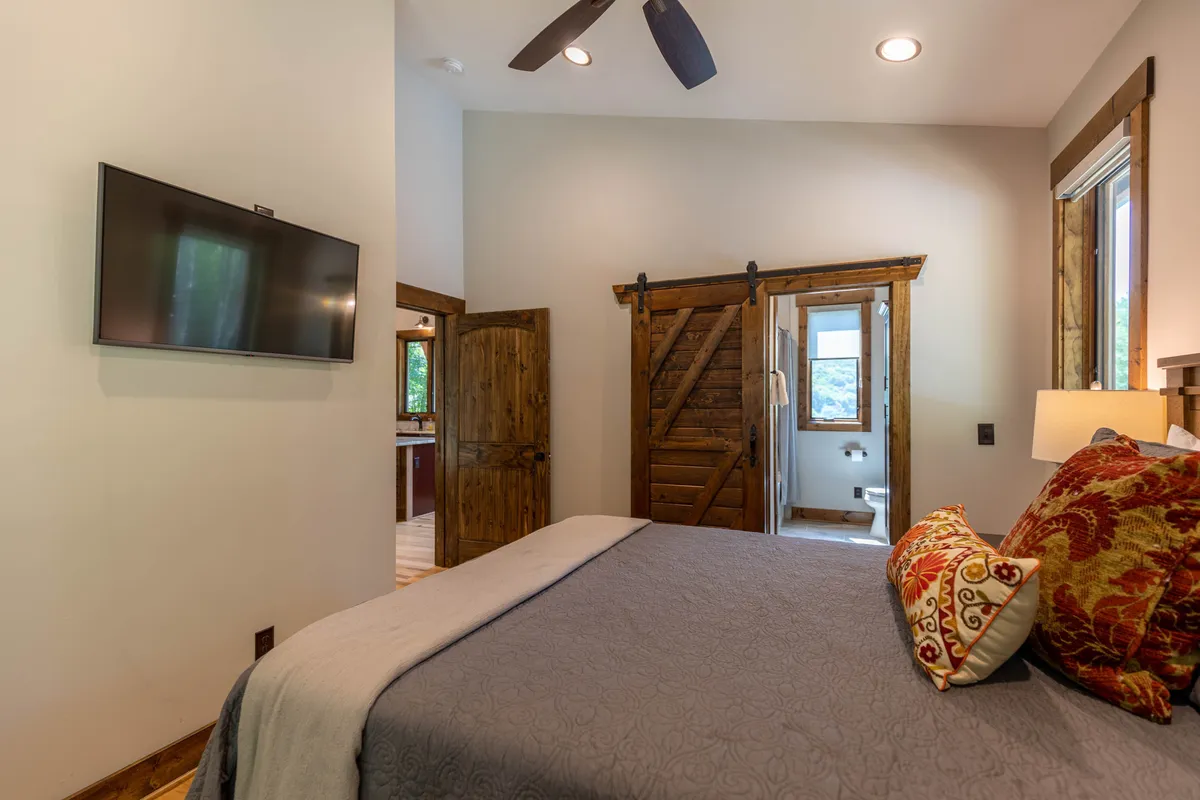 This interior shot features a bed with decorative pillows, a sliding barn door, and a wall-mounted television. Natural light filters gently through the window, and the design elements create a comfortable, inviting space perfect for unwinding with a movie or a book.