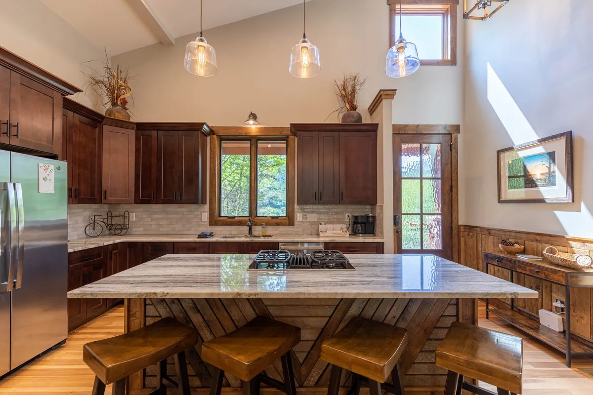 The image showcases a cozy kitchen with a central island and bar stools, framed by a large window overlooking lush greenery. Warm wooden cabinetry and inviting decor create a welcoming atmosphere, while natural light streams in, highlighting the space’s connection to the outdoors.