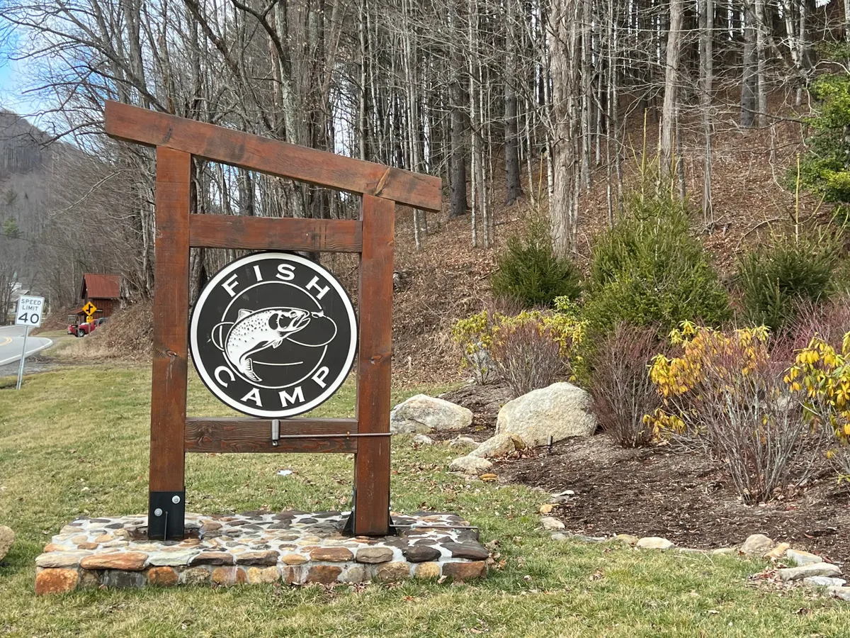 A relaxed, landscaped area centered around a wooden sign with a fish emblem, hinting at fishing activities. The setting conveys both a sense of arrival and the promise of outdoor adventures such as fishing and camping.