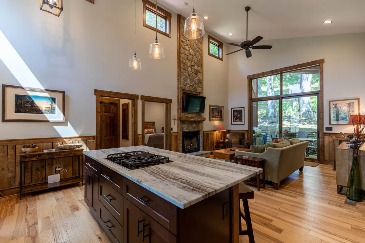 A bright, airy interior with a kitchen island featuring a stove, a stone fireplace, large windows allowing for abundant natural light and scenic views, and a comfortable seating area. The design encourages group cooking, conversation, and relaxation.