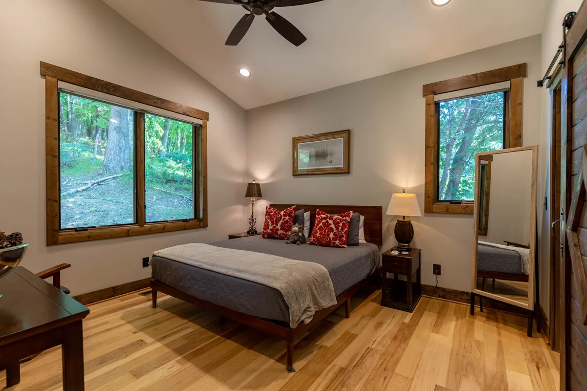 A bright bedroom with a queen-sized bed, large windows offering views of the greenery outside, and inviting wooden furniture. The tranquil setting visually connects the comfort of the indoors with the beauty and serenity of the outdoors.