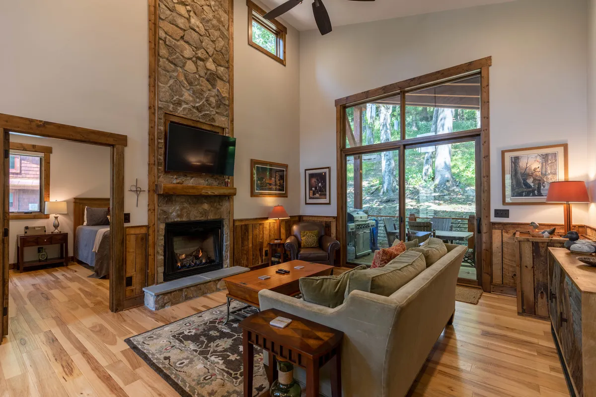 This image showcases a cozy interior space anchored by a stone fireplace, large windows that invite natural light, and rustic wooden furniture. People are seen relaxing by the fire, socializing, and reading, creating a welcoming and communal atmosphere.