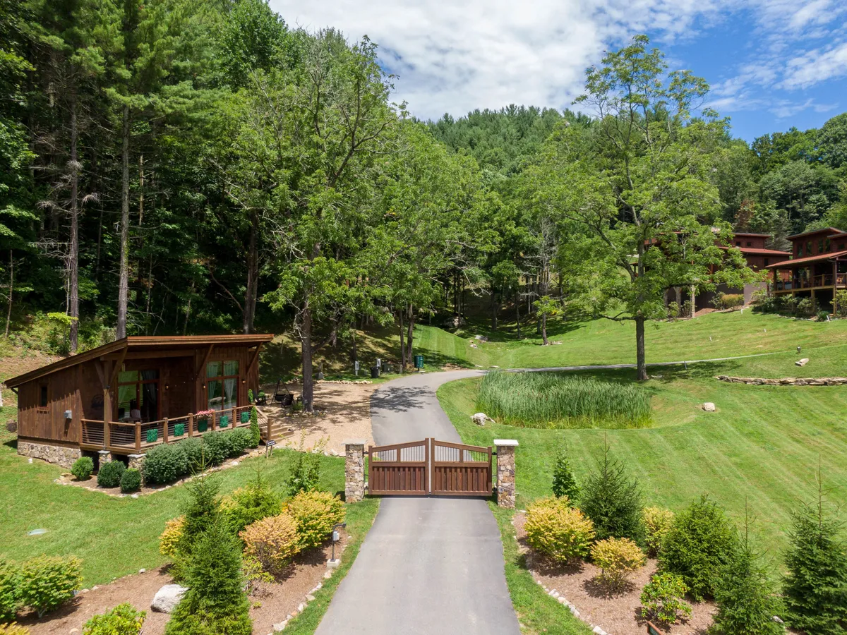 This image captures the wooden chalet exterior framed by lush greenery, with a prominent gated entrance and a driveway welcoming guests into the serene mountain environment. The porch and landscaped grounds suggest ample space for outdoor activities, gatherings, and pet-friendly exploration.