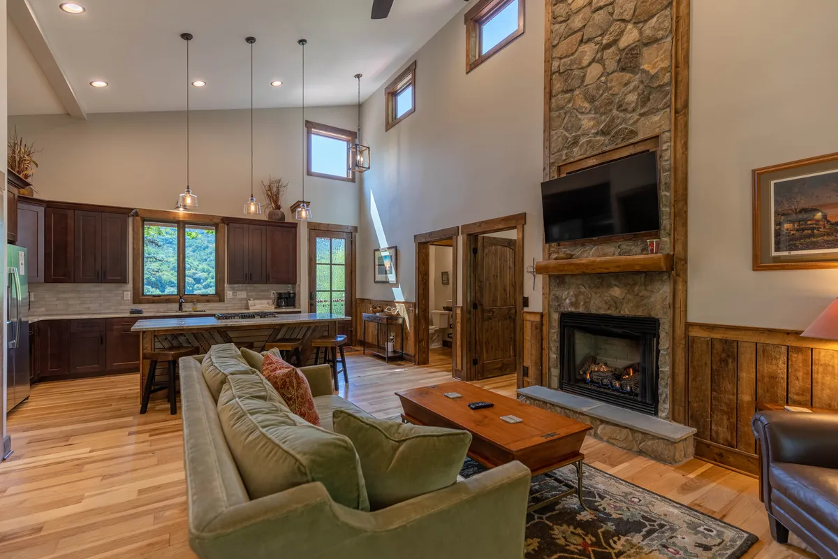A spacious open-concept living area that seamlessly joins the living room, kitchen, and dining spaces. The room features a striking stone fireplace, a substantial kitchen island with seating, large windows that bring in mountain views, a comfortable sofa, and a dining table, all under warm, inviting lighting.