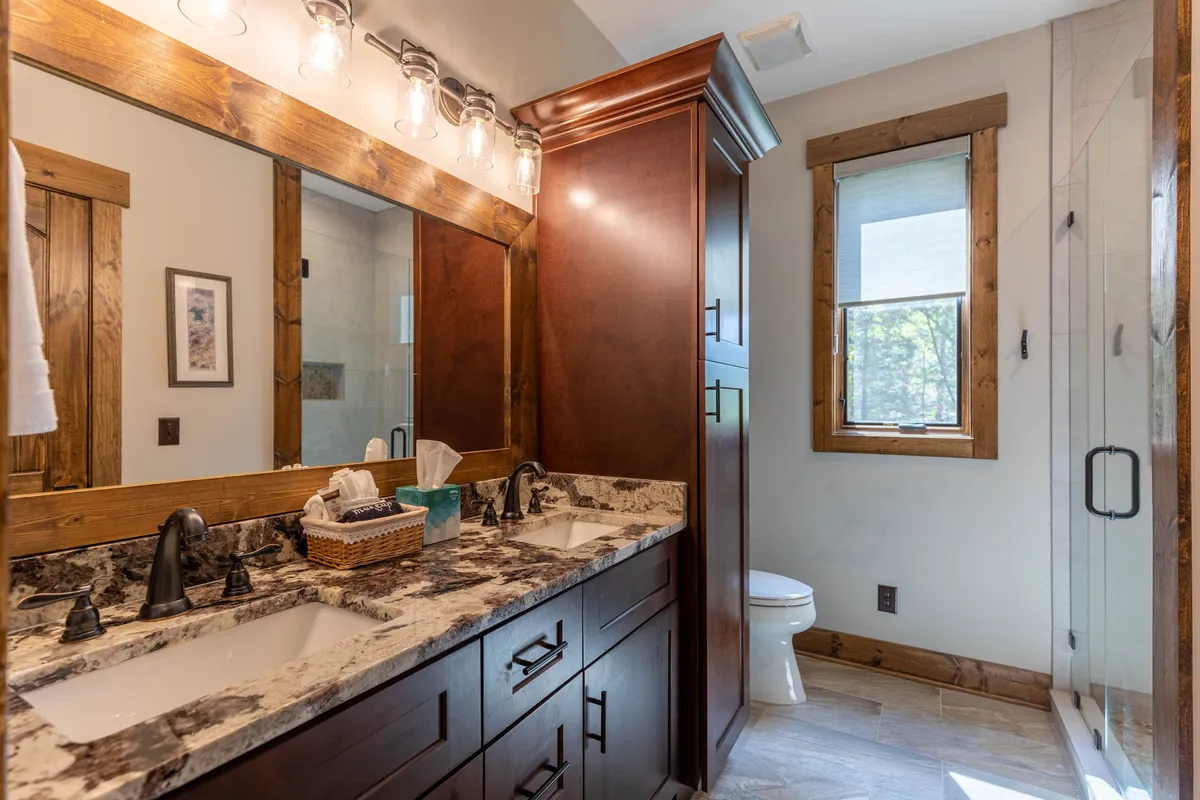 A double sink vanity with a large mirror and wooden cabinetry is showcased in this calming bathroom space. The shower area is also visible, and the overall design supports a sense of luxury and self-care.