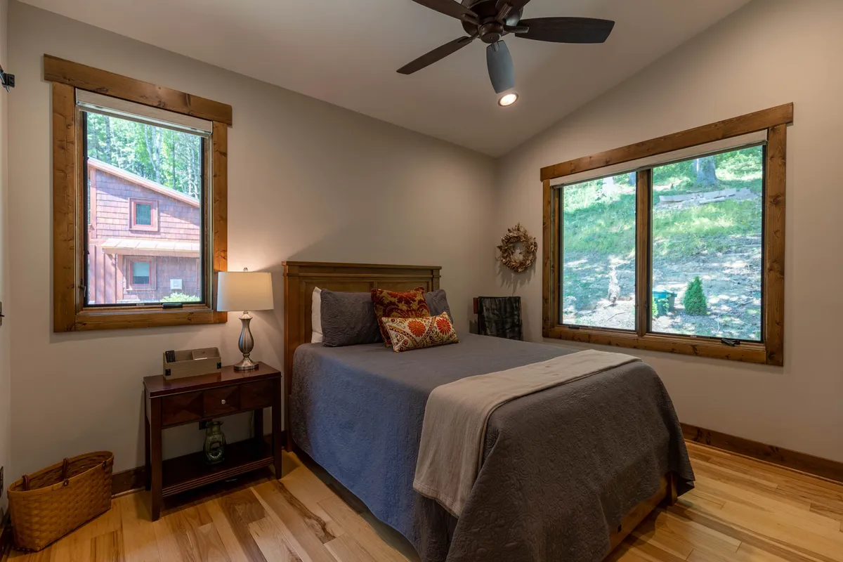 This image shows a tranquil bedroom with a comfortable bed adorned with decorative pillows, a nightstand with a lamp, and large windows letting in soft natural light. The view outside offers a calming connection to nature, and the overall mood is restful and serene.